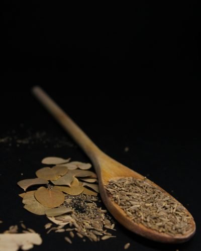 Close-up of cumin seeds and bay leaves on a wooden spoon against a dark background.
