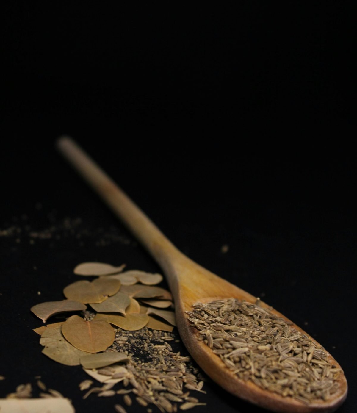 pexels-photo-5774814-5774814 Close-up of cumin seeds and bay leaves on a wooden spoon against a dark background.